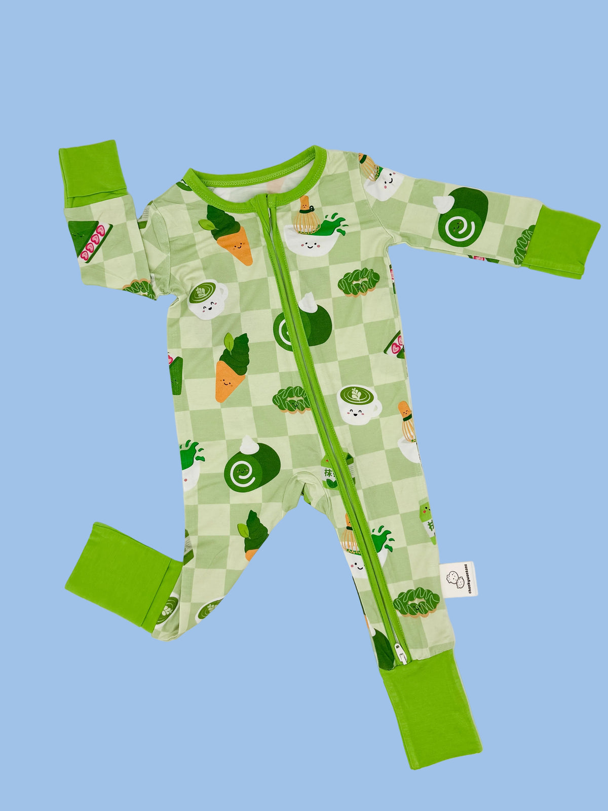A baby long-sleeve footed sleeper with a green and white checkered pattern, featuring cartoon vegetables like carrots, broccoli, and peas, with solid green cuffs and a green zipper.